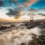 Weißer Windmühlenbau auf felsigem Ufer bei Sonnenuntergang mit welligem Meer.