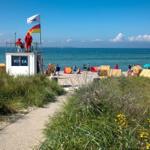 Strand mit Rettungswache, Liegestühlen und Menschen. Pfad durch Gras zum Wasser.
