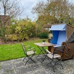 Terrasse mit Tisch, zwei Stühlen und Strandkorb. Grünfläche und Bäume im Hintergrund.