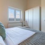 Schlafzimmer mit Bett, Kleiderschrank und Fenster. Tür und Stuhl sind sichtbar.