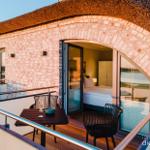 Terrasse mit Tisch und Stühlen. Durch Glastür ist Schlafzimmer mit Bett sichtbar.