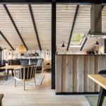 Living and kitchen area with wooden wall and ceiling. Dining table, sofa, and windows.
