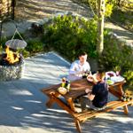 Zwei Personen sitzen an einem Holztisch im Garten und grillen an einem Feuerkessel.