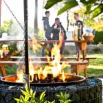 Ein brennender Feuerkorb mit Holz im Garten. Im Hintergrund steht ein Holztisch mit Personen.