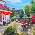 Terrasse mit rotem Sonnenschirm und Gartenmöbeln. Holzdielen und Pflanzen umgeben die Loungezone.
