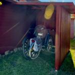 Zwei Fahrräder stehen unter einem roten Carport. Gras und Steinmauer sind sichtbar.