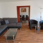 Wohnzimmer mit Sofa, zwei Sesseln und Glas-Tisch. Durchgang zur Essnische.