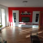 Wohnzimmer mit roter Wand, weißen Schränken und TV. Holzboden und Glas-Tisch.