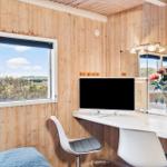 A room with wooden walls, desk, monitor, and bed. Window shows landscape.