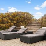 Three lounge chairs with cushions are on a wooden terrace in front of shrubs.
