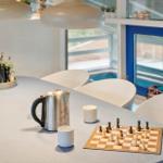 Table with chessboard, kettle, and cups. White chairs and large windows.