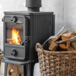 Schwarzer Holzofen mit brennendem Feuer und Korb mit Brennholz neben einem rustikalen Stuhl.