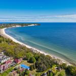 Aerial view of a beach with coastal houses and greenery under a clear blue sky.
