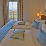 Zwei Betten mit Kissen und Decke. Fenster zum Balkon mit Meerblick. Wandbild und Fernseher.