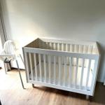 Kinderzimmer mit Hochstuhl und Babybett. Holzboden und Fenster mit Vorhang.