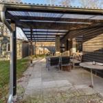 Terrace with pergola, tables and chairs beside wooden house. Lawn area and small outbuilding in background.