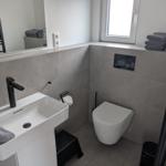 Badezimmer mit Waschbecken, Toilette und Fenster. Graue Fliesen und schwarze Accessoires.