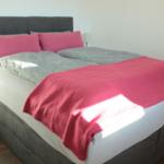 Schlafzimmer mit Doppelbett, rosa Kissen und Decke, grauer Kopfteil und Holzboden.