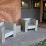 Zwei hölzerne Sitzgelegenheiten stehen auf einem gepflasterten Hof vor einer Backsteinmauer.