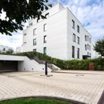 Modernes weißes Gebäude mit gepflastertem Vorplatz und Treppen zur Eingangsebene.