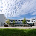 Moderne weiße Gebäude mit Glasfassade und grüner Umgebung unter blauem Himmel.