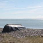A black boat half lies on the pebble beach. Water stretches in the background.