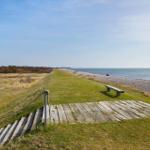 Wooden path leads to the beach. Grass area, bench, and view of the sea.