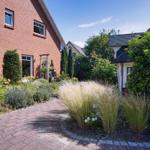 Gartenweg mit Blumenbeeten und hohem Gras vor einem Backsteinhaus.