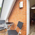 Terrasse mit Tisch und zwei Stühlen neben roter Ziegelwand. Blick ins Innere mit Holzboden.