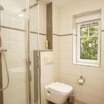 Badezimmer mit Dusche, Toilette und Fenster mit Blick auf Bäume.