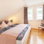 Schlafzimmer mit Bett, Stuhl und zwei Fenstern mit Vorhängen.