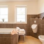 Badezimmer mit Badewanne, Toilette und Waschbecken. Zwei Fenster und braune Fliesen.