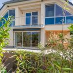 Modernes Haus mit Balkon und Terrasse, umgeben von Grün und einem kleinen Teich.