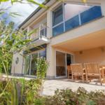 Haus mit Terrasse, Garten und Holzmöbel. Blick auf Pool und Bäume.