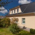 Ein Haus mit braunem Dach und hellen Wänden, umgeben von Grünflächen und Sträuchern.