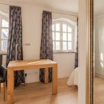 Ein Zimmer mit Schreibtisch, Stuhl und Fenstern. Spiegel zeigt Kleiderschrank.