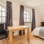 Schlafzimmer mit Bett, Schreibtisch und zwei Fenstern mit Vorhängen.