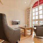 Zwei graue Sessel und ein Holztisch stehen vor einem großen Fenster mit roten Jalousien.