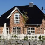 Rotes Backsteinhaus mit schwarzem Dach und weißem Zaun. Garten mit Steinen und Pflanzen.