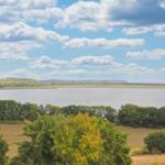 Weitläufiger See mit kleinem Boot, umgeben von Wiesen und Bäumen unter blauem Himmel mit weißen Wolken.
