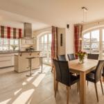 Küche und Essbereich mit Holztisch, Stühlen und Kaminofen. Große Fenster mit Blick nach draußen.