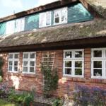 Haus mit strohgedecktem Dach und roten Ziegelmauern, weiße Fenster und Blumenbeete.