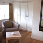 Zimmer mit Sitzgelegenheit, Kleiderschrank und Spiegel. Fenster mit Vorhängen.