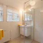 Badezimmer mit Dusche, Waschbecken und zwei Fenstern mit Jalousien.