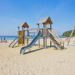 Strandspielplatz mit Rutsche und Schaukel. Sandstrand und ruhiges Wasser im Hintergrund.