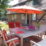 Terrasse mit Tisch, Stühlen und orangefarbenem Sonnenschirm vor einem Haus mit Strohdach.