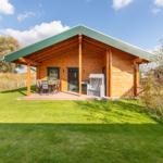 Holzhaus mit Terrasse, Garten und Liegestuhl unter blauem Himmel.
