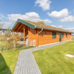 Holzhaus mit grünem Dach, Terrasse und Rasenfläche. Pfad führt zur Eingangstür.