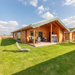 Holzhütte mit Terrasse, Garten und Liegestuhl unter blauem Himmel.