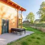 Haus mit Holzfassade und Terrasse mit Tisch und Stühlen. Grünfläche und Zaun im Hintergrund.